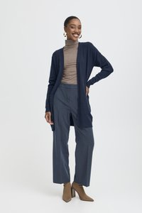 Mosolygó nő arany karika fülbevalóval, barna garbóval, tengerészkék kardigánnal, kék bő szárú nadrággal és bézs magassarkú bokacsizmával, kezét a csípőjén tartva áll.