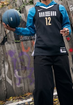 Person trägt schwarzes Basketballtrikot Nummer 12 und blauen Hoodie und hält einen blauen Basketball neben einem Holzzaun mit Graffiti.