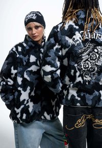 Luddig, svart och grå kamouflagejacka med dragkedja, hög krage och ett broderat drakmotiv på ryggen. Modellen bär en svart mössa.