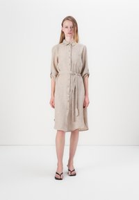 Robe chemise en lin beige avec un col, un devant boutonné et une taille à nouer. Dotée de manches retroussées et d'une poche poitrine. Portée avec des sandales noires.