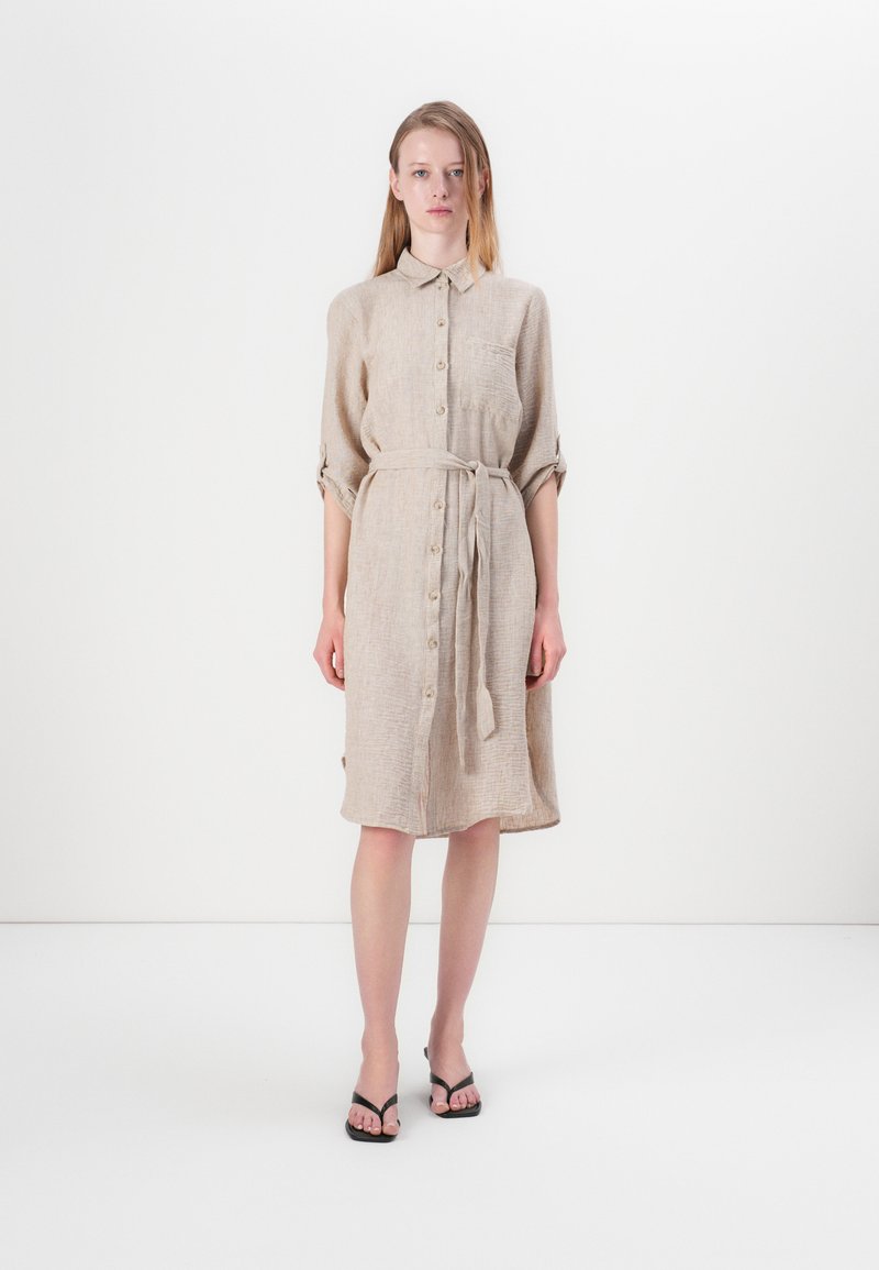 Robe chemise en lin beige avec un col, un devant boutonné et une taille à nouer. Dotée de manches retroussées et d'une poche poitrine. Portée avec des sandales noires.