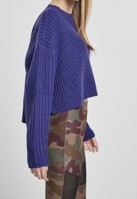 Lila stickad tröja med lös passform och ribbad struktur, ihop med kamouflagemönstrade leggings i bruna, gröna och svarta nyanser.