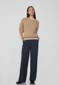 Beige Kabelstrickpullover mit kurzen Ärmeln, über einem weißen Hemd getragen. Marineblaue Hellleg-Jeans aus strukturiertem Stoff, mit schwarzen Schuhen.