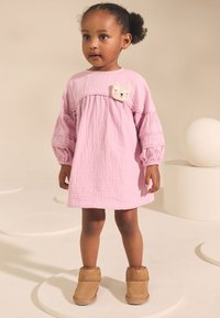 Robe en coton rose avec manches bouffantes et un patch chat à motifs. Associée à des bottines beiges en peluche sur un fond uni.