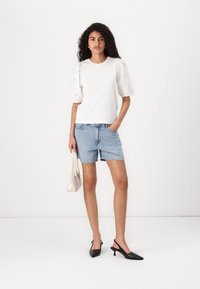 Vit kortärmad topp med puffärmar, denimshorts och svarta slingback-klackar. Håller en liten krämfärgad handväska. Slätt tyg, avslappnad design.