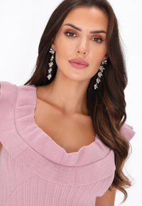 Roze gebreide top met een gerimpelde halslijn, met een textuur afwerking; model draagt hangende bloemenoorbellen met parels en accenten.