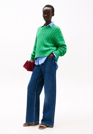 Grøn strikket sweater med ribbet tekstur over en blå stribet skjorte, parret med løse mørkeblå jeans og sandaler med åben tå. Rød håndtaske.