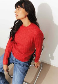 Pull en tricot rouge à côtes avec un motif texturé et un col rond, associé à un jean en denim bleu taille haute. Assis sur une chaise métallique.