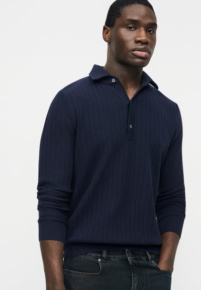 Homme portant un pull côtelé à manches longues bleu marine avec un col partiellement boutonné et un jean foncé, debout devant un fond uni.