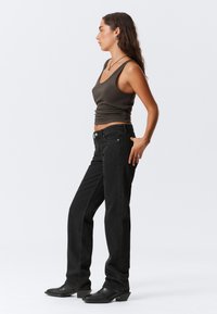 Pikkade juustega naine, kes kannab tumedat topist, musti sirgeid teksaseid, musti kontsakingi ja hõbedaseid ehteid, seistes külg ees valge tausta ees.