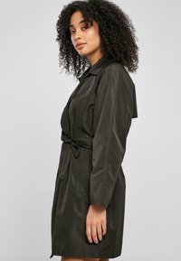 Trench coat ușor, de culoare verde închis, cu talie accesorizată cu un brâu, mâneci lungi și buzunare laterale. Textură netedă, design până la genunchi.