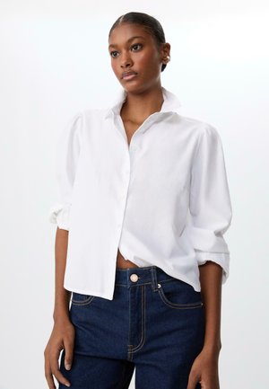 Camicetta bianca con maniche arrotolate e colletto, realizzata in tessuto leggero, abbinata a jeans blu scuro con cuciture dorate.