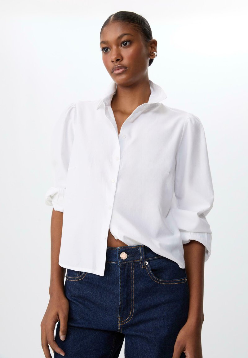 Chemise blanche à manches retroussées et col, fabriquée en tissu léger, associée à un jean bleu foncé avec des coutures dorées.