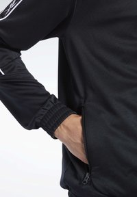 Černá sportovní bunda s kapsou na zip, elastickými manžetami a hladkým materiálem. Jemné bílé akcenty podél ramene.