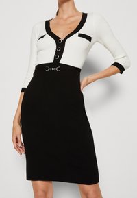 Stickad klänning med vit överdel och svart kjol. Har en V-hals, tre knappar, svarta detaljer och ett svart bälte med en silverspänne.