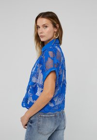 Blue mesh blouse with floral lace pattern and short sleeves. Features a collared neckline and scalloped hem. Worn over light denim jeans.