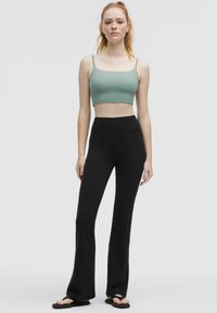Grünes geripptes Crop-Top mit dünnen Trägern, kombiniert mit hochgeschnittenen schwarzen Schlaghosen und schwarzen Flip-Flops vor einem neutralen Hintergrund.