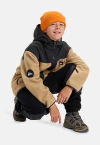 Giacca per ragazzi con parte superiore in nylon nero, parte inferiore in felpa beige, tasche con zip e patch con logo. Abbinata a pantaloni sportivi scuri e sneakers con texture.