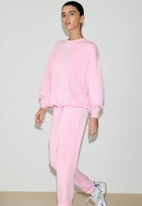 Svetloružová mikina a zodpovedajúce tepláky z jemnej látky, s voľným strihom, spustenými ramenami a manžetovými rukávmi.