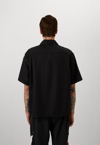 Black short-sleeve shirt with a collar, featuring a relaxed fit. Back design includes a yoke, minimal stitching, and soft fabric texture.