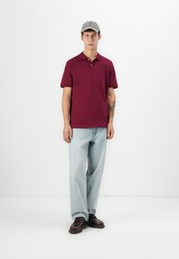 Bordeauxfarvet polo skjorte med krave, båret med lyseblå jeans og brune snørresko. En grå kasket fuldender outfittet, med en tatovering synlig på armen.