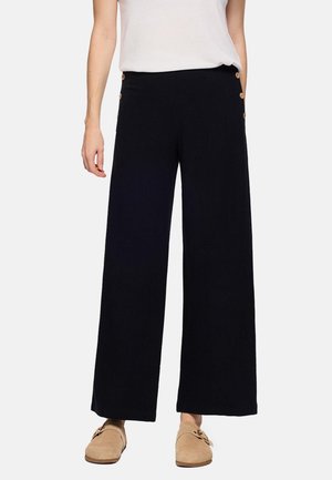 Mujer vestida con pantalones negros de pierna ancha con botones laterales y zapatos beige sin cordones, combinados con una blusa blanca sin mangas.