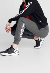 Zwarte jas met ritssluiting en drie witte strepen, rood T-shirt, grijze joggingbroek met wit "adidas"-logo en zwarte sportschoenen met witte accenten.