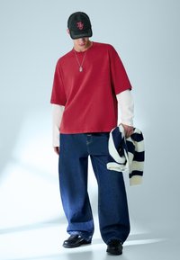 Mladý muž v oversized červené košili s krátkými rukávy na bílé košili s dlouhými rukávy, široké modré džíny, černá čepice, černé boty, drží pruhovaný svetr.