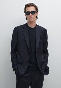 Dunkel gestreifter Blazer über einem schwarzen Rundhals-T-Shirt, kombiniert mit rechteckigen schwarzen Sonnenbrillen, schmaler Schnitt und glatte Textur.
