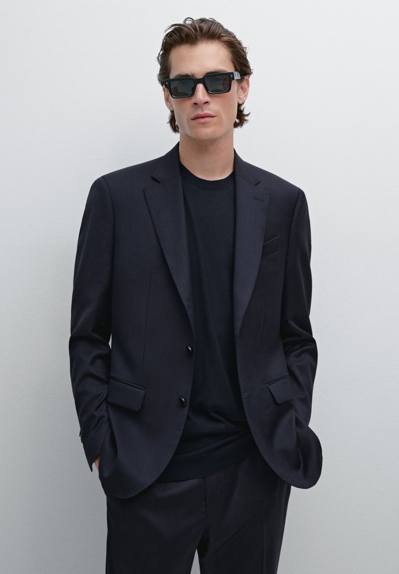Dunkel gestreifter Blazer über einem schwarzen Rundhals-T-Shirt, kombiniert mit rechteckigen schwarzen Sonnenbrillen, schmaler Schnitt und glatte Textur.