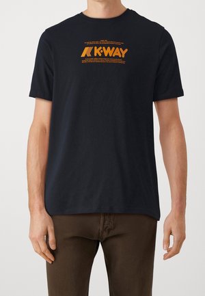 Man wearing black short-sleeve T-shirt with orange "K-WAY" logo and text centered on chest, paired with dark brown pants.