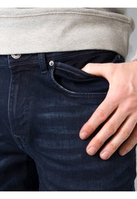 Jeans de mezclilla azul oscuro con un acabado texturizado, que presentan un bolsillo frontal y un aspecto ligeramente desgastado. La mano está colocada en el bolsillo.