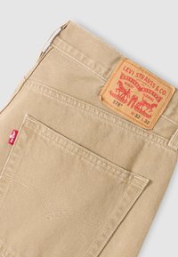 Μπεζ βαμβακερό παντελόνι twill με δερμάτινο πίσω ταμπελάκι που φέρει ανάγλυφο λογότυπο της μάρκας και δύο πίσω τσέπες· χαρακτηριστική κόκκινη ετικέτα στην αριστερή τσέπη.