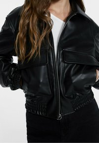 Veste bomber en cuir noir avec une fermeture éclair à l'avant, deux poches sur la poitrine et des poignets en côtes. Le matériau semble lisse avec une coupe ajustée.