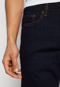 Jeans de denim azul oscuro con costuras naranjas, que presentan un bolsillo frontal y una mano descansando cerca de la cintura. La tela parece suave con un ligero brillo.