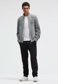 Graue Jacke mit Reißverschluss und gerippten Ärmeln, mit Grafikprints, kombiniert mit einem weißen T-Shirt und schwarzen Hosen. Die Schuhe sind weiß mit hellgrauen Akzenten.