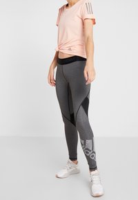 Světle růžové sportovní tričko s uzlem vpředu, šedé legíny s černými a síťovanými akcenty a bílé tenisky se šedými detaily.