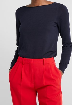 Femme portant un haut bleu marine à manches longues rentré dans un pantalon plissé rouge vif taille haute, les mains dans les poches.