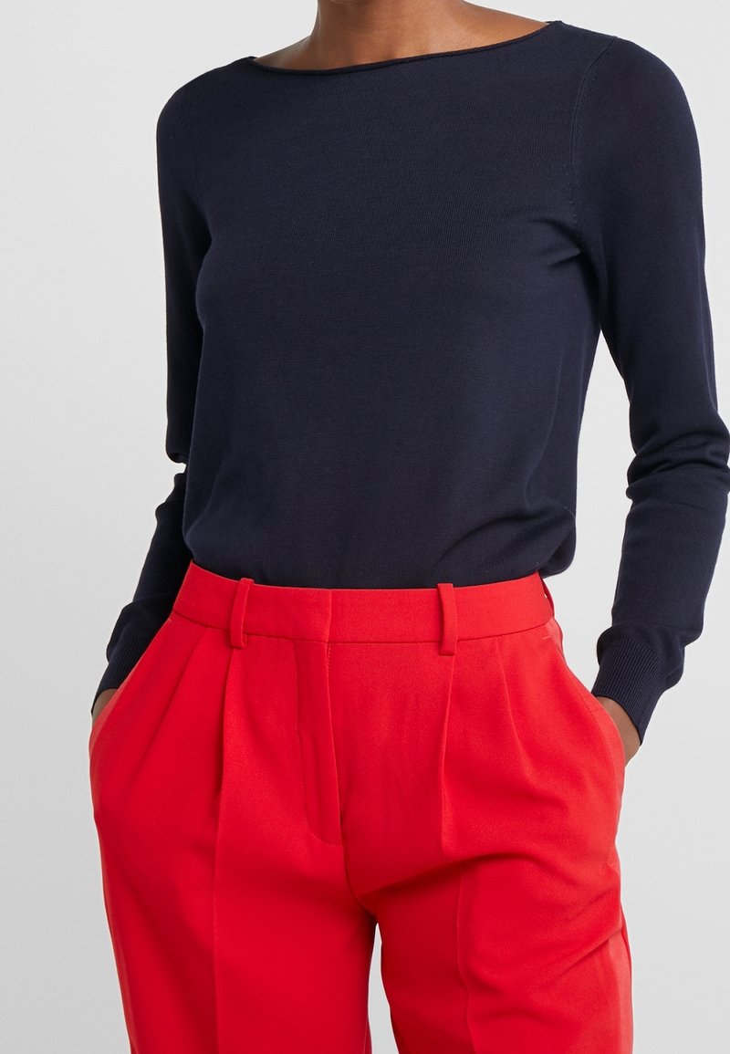 Femme portant un haut bleu marine à manches longues rentré dans un pantalon plissé rouge vif taille haute, les mains dans les poches.