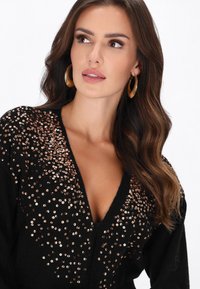 Frau mit langen, welligen braunen Haaren, die große goldene Creolen und einen schwarzen V-Ausschnitt-Pullover mit goldenen Pailletten trägt.