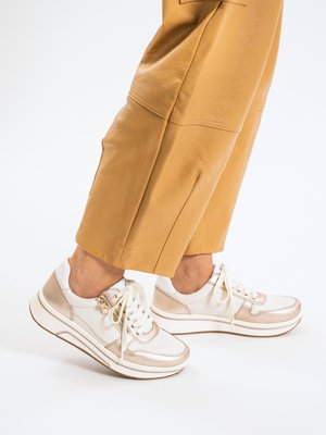 Gambe con pantaloni corti color sabbia e sneakers bianche con dettagli in oro rosa e lacci bianchi su sfondo bianco.