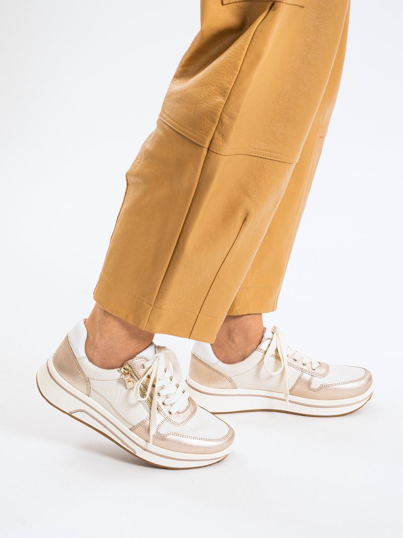 Legs wearing tan cropped pants and white sneakers with rose gold accents and white laces on a white background.