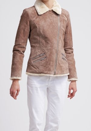 Mujer con chaqueta ajustada de ante marrón con cuello y puños de piel crema, cremallera frontal y dos bolsillos con cremallera, combinada con pantalones blancos.