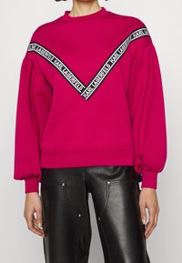 Fuksjowa bluza z długimi, bufkami rękawami i okrągłym dekoltem. Posiada czarne logo w kształcie litery V na piersi.