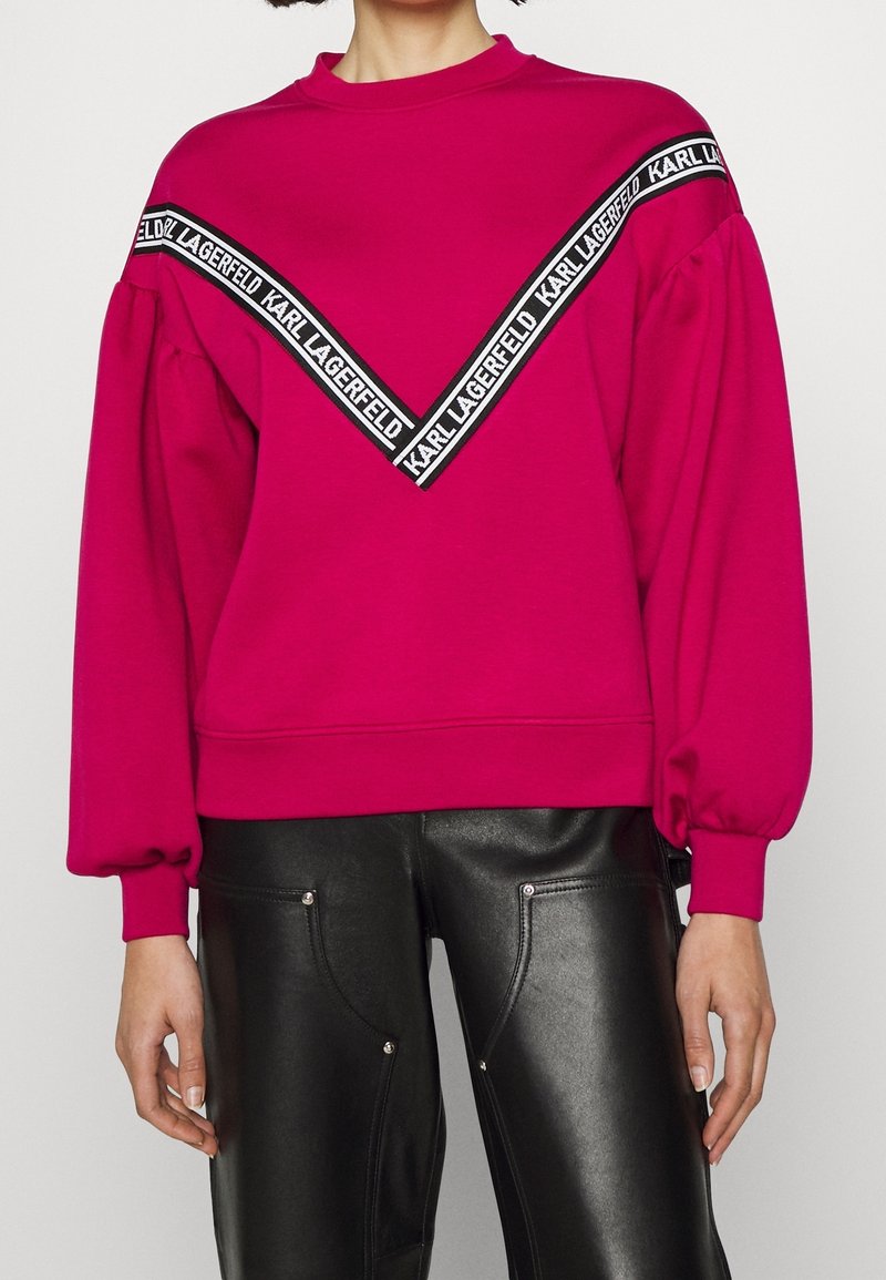 Fuksjowa bluza z długimi, bufkami rękawami i okrągłym dekoltem. Posiada czarne logo w kształcie litery V na piersi.