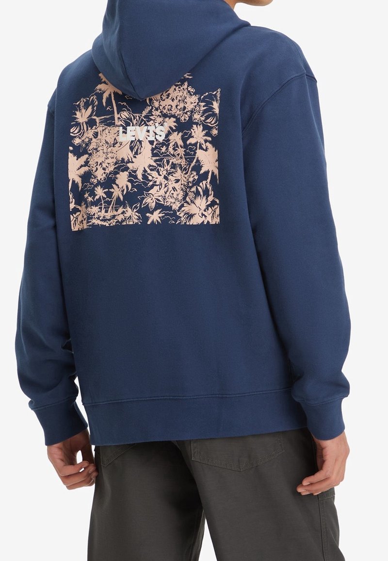 Personne portant un sweat à capuche bleu marine avec un imprimé floral tropical beige et le logo "LEVI'S" au dos, assorti à un pantalon foncé.