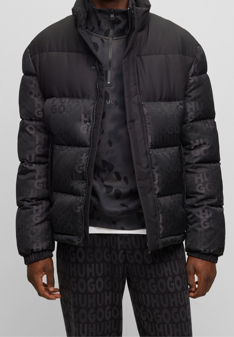 Homme portant une veste matelassée noire par-dessus un haut zippé noir à motifs et un pantalon assorti avec le texte "HUGO" répété.