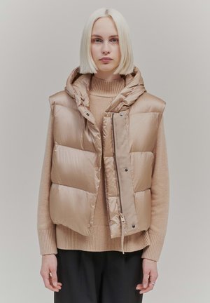Padded sleeveless vest in light beige, featuring a hood, zip closure, and snap buttons, layered over a beige sweater and paired with dark pants.