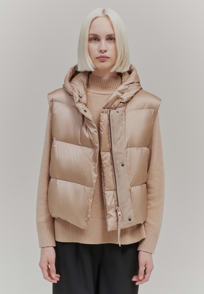 Padded sleeveless vest in light beige, featuring a hood, zip closure, and snap buttons, layered over a beige sweater and paired with dark pants.