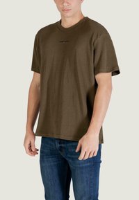 T-shirt verde oliva in cotone con scollo rotondo, maniche corte e un piccolo logo "TOMMY JEANS" sul petto. Indossata con jeans blu.